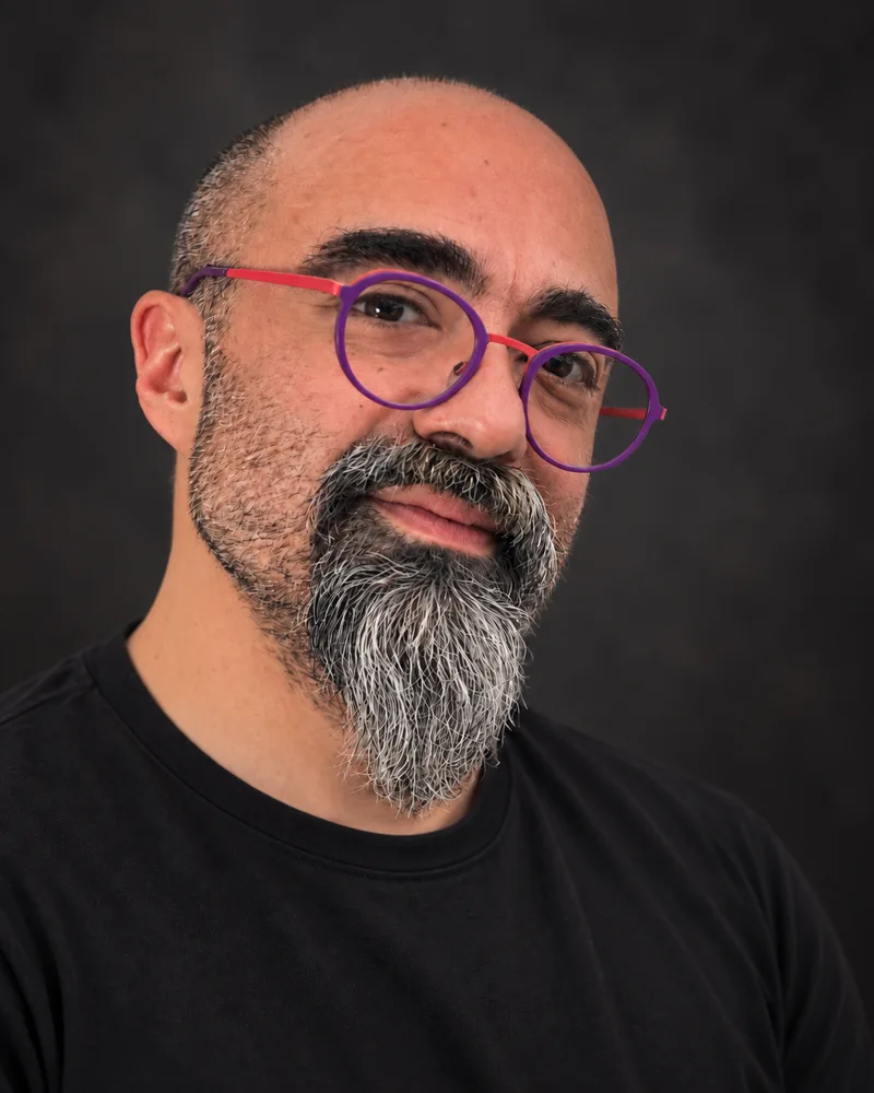 Onur, wearing distinctive purple glasses and a black shirt, seated beside a window.